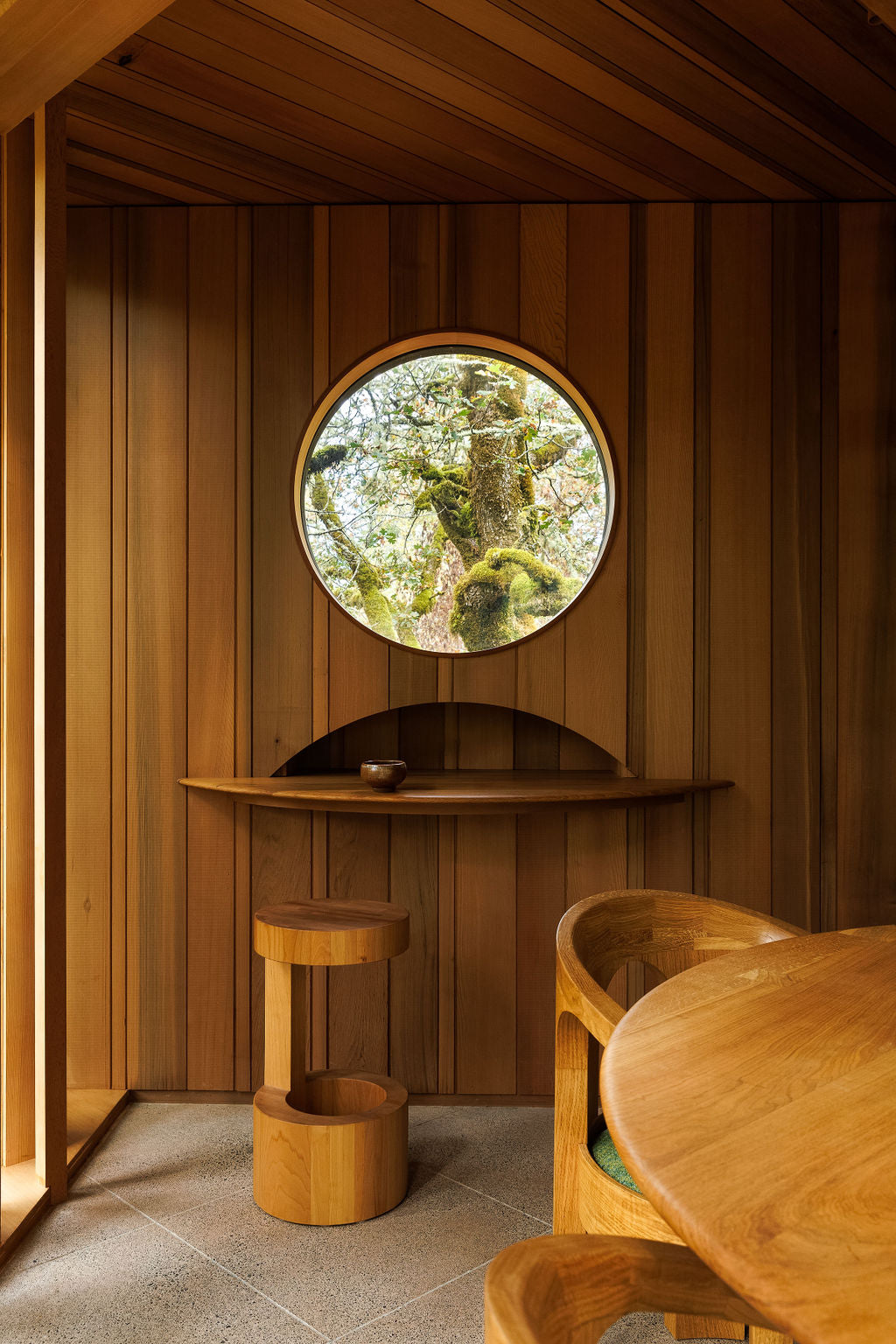 A handmade side table, built-in wall shelf and dining table and chairs in a wood-panelled room. All of the wood matches with a warm chestnut brown colour. 