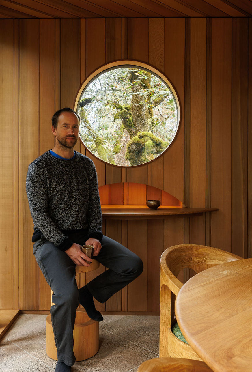 Eric Gesinger sits in a room filled with wood paneling and wooden furniture made by him. All of the wood matches with a warm chestnut brown colour