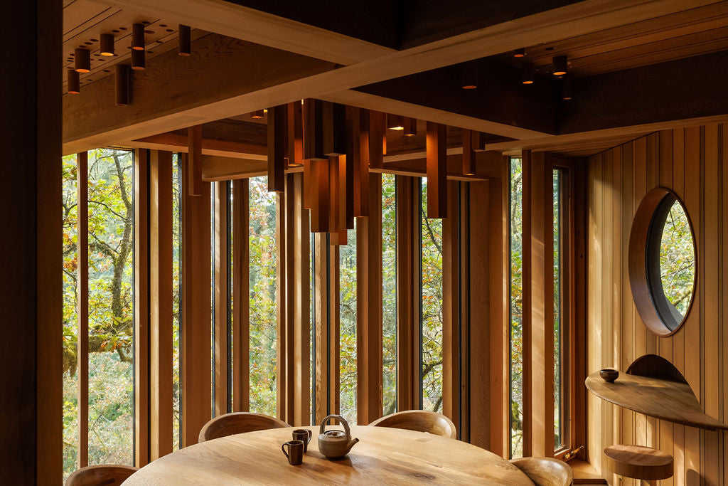 A room filled with wood paneling and handmade wooden furniture. All of the wood matches with a warm chestnut brown colour. And light spills in from the vertical windows showing a green canopy outside.