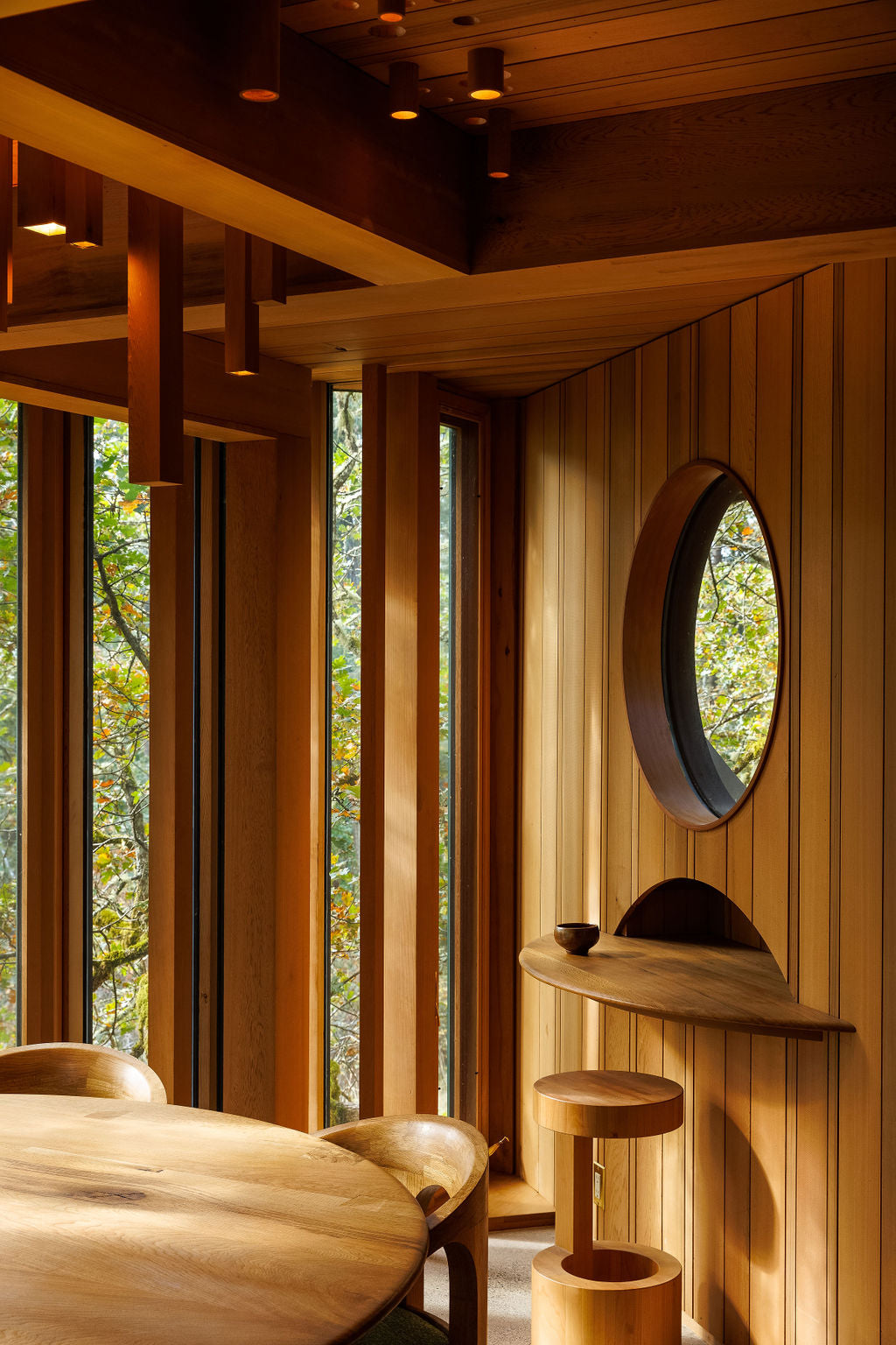 A room filled with wood paneling and handmade wooden furniture. All of the wood matches with a warm chestnut brown colour. And light spills in from the vertical windows showing a green canopy outside.