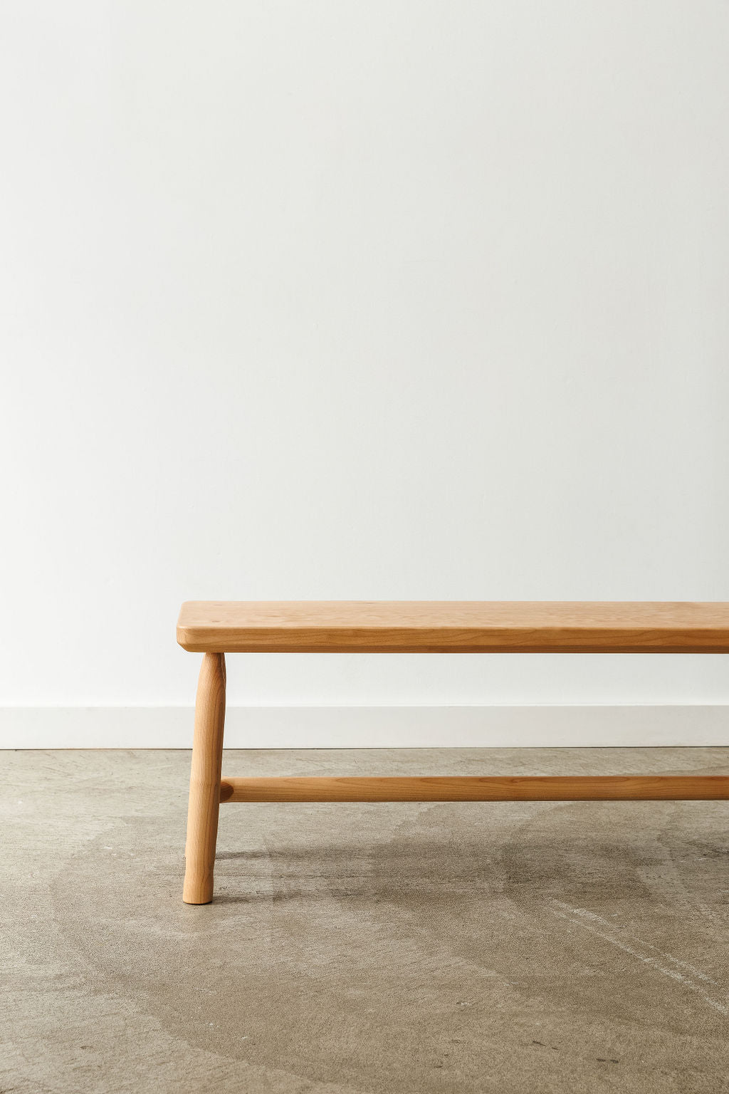 A light coloured wooden bench in a white room with concrete floors. Showing it's front profile.