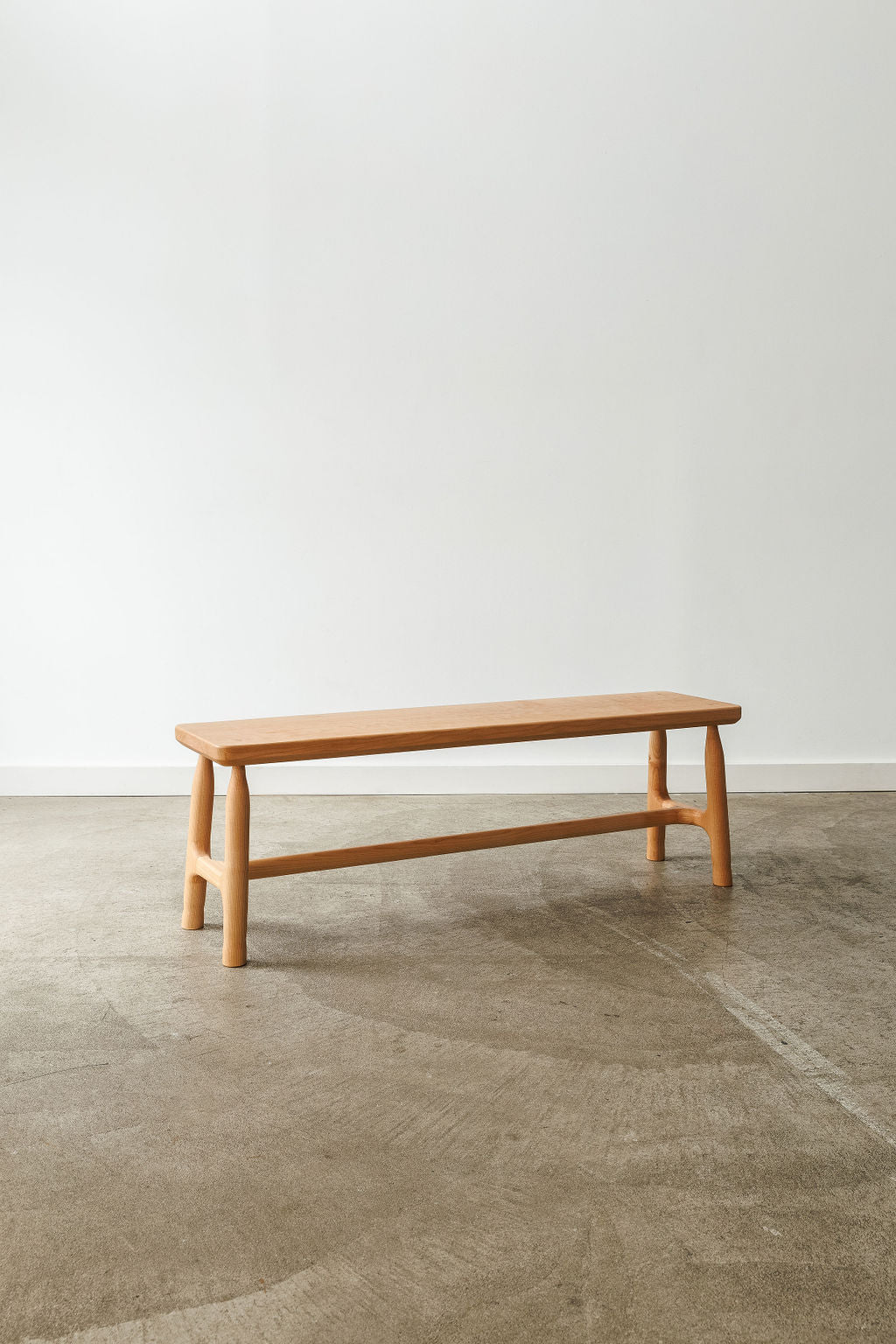 A light coloured wooden bench in a white room with concrete floors. Showing it's front profile.