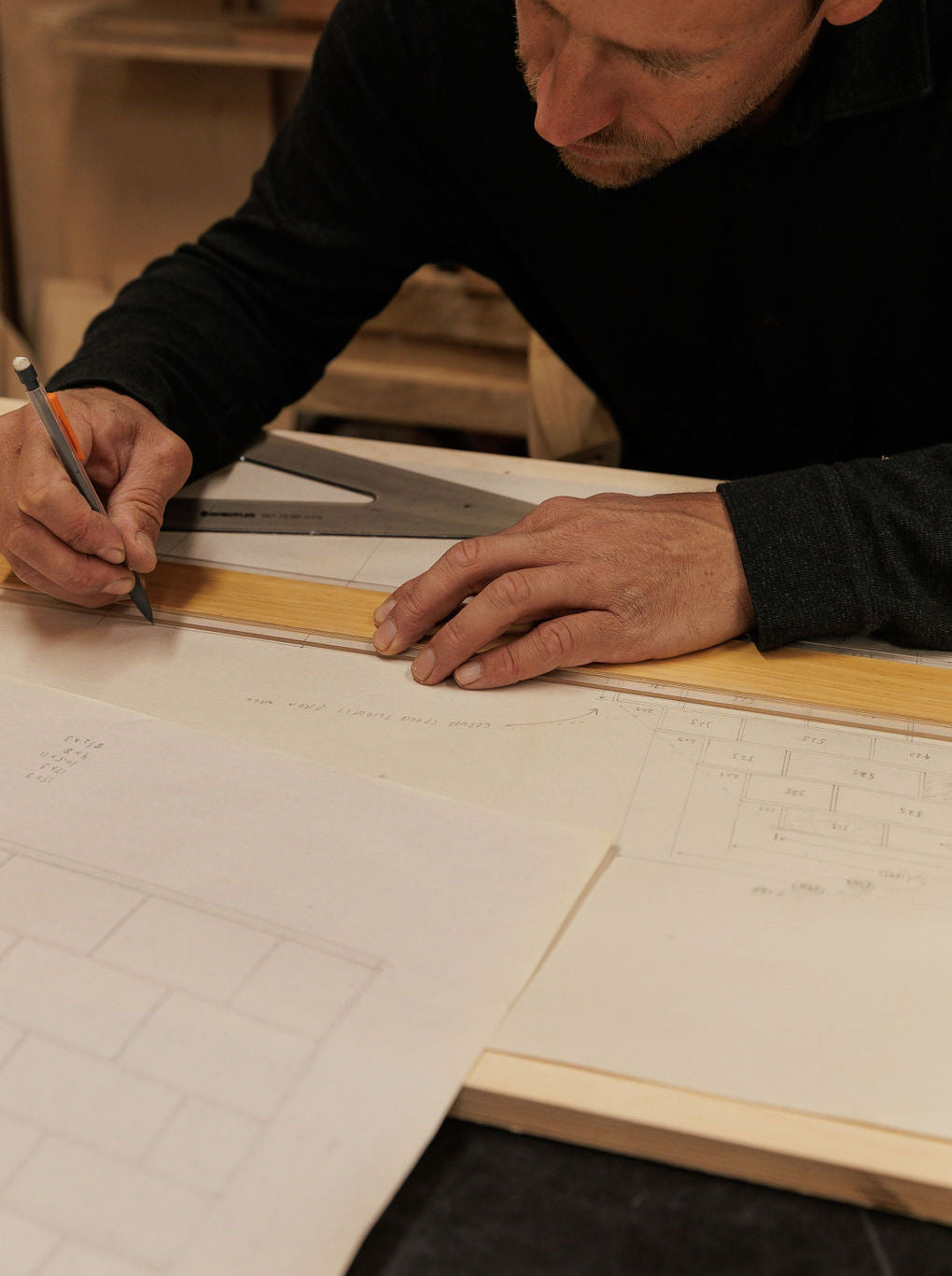 Eric is sitting at a drafting table in his shop bent over a plan holding a pencil and ruler.