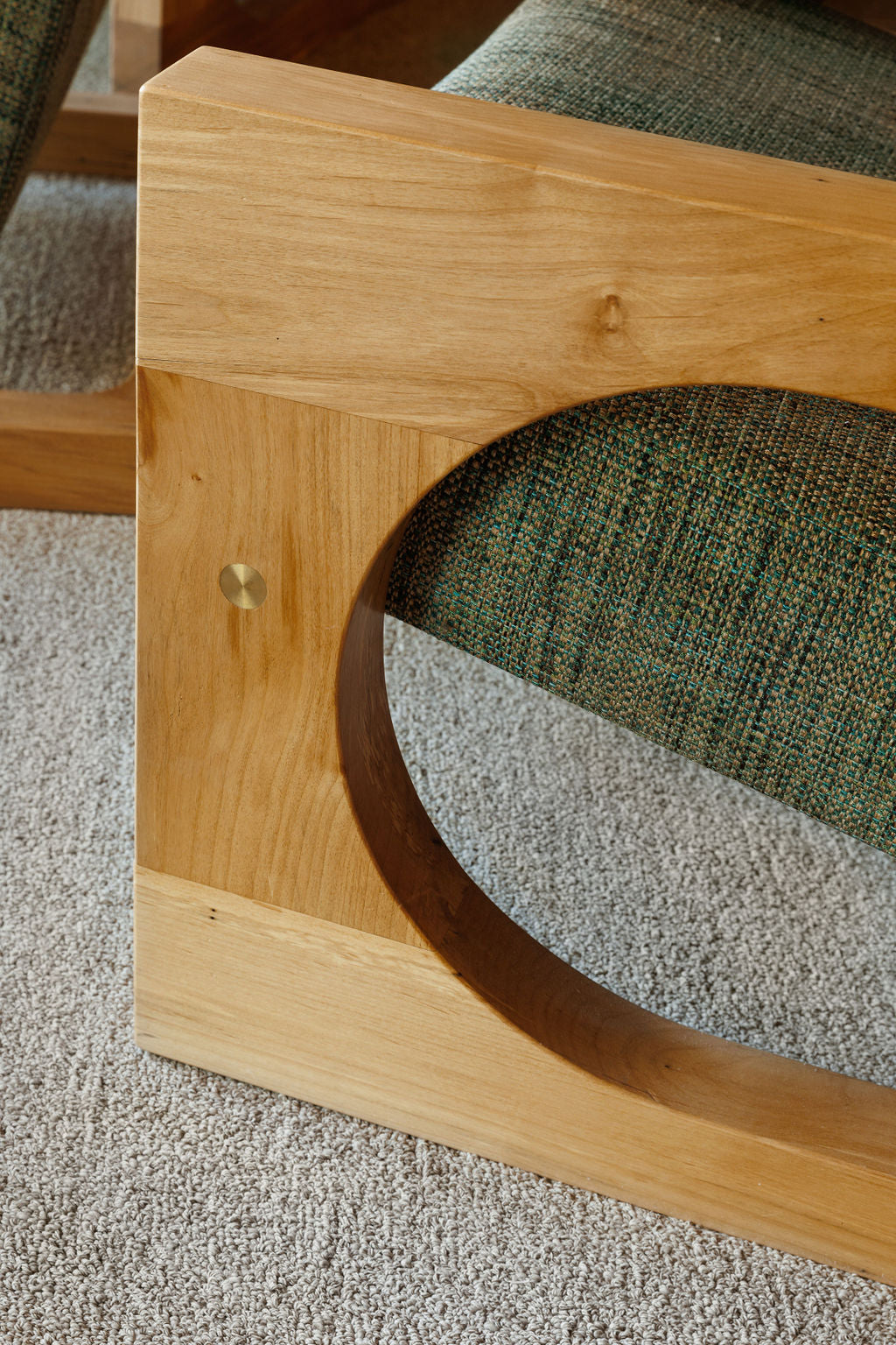 The side of a custom wood arm chair is shown. The arm is a large rectangle with a large circle cut out of the middle to create the arm and bottom foot of the chair. The chair is in a warm medium toned wood.