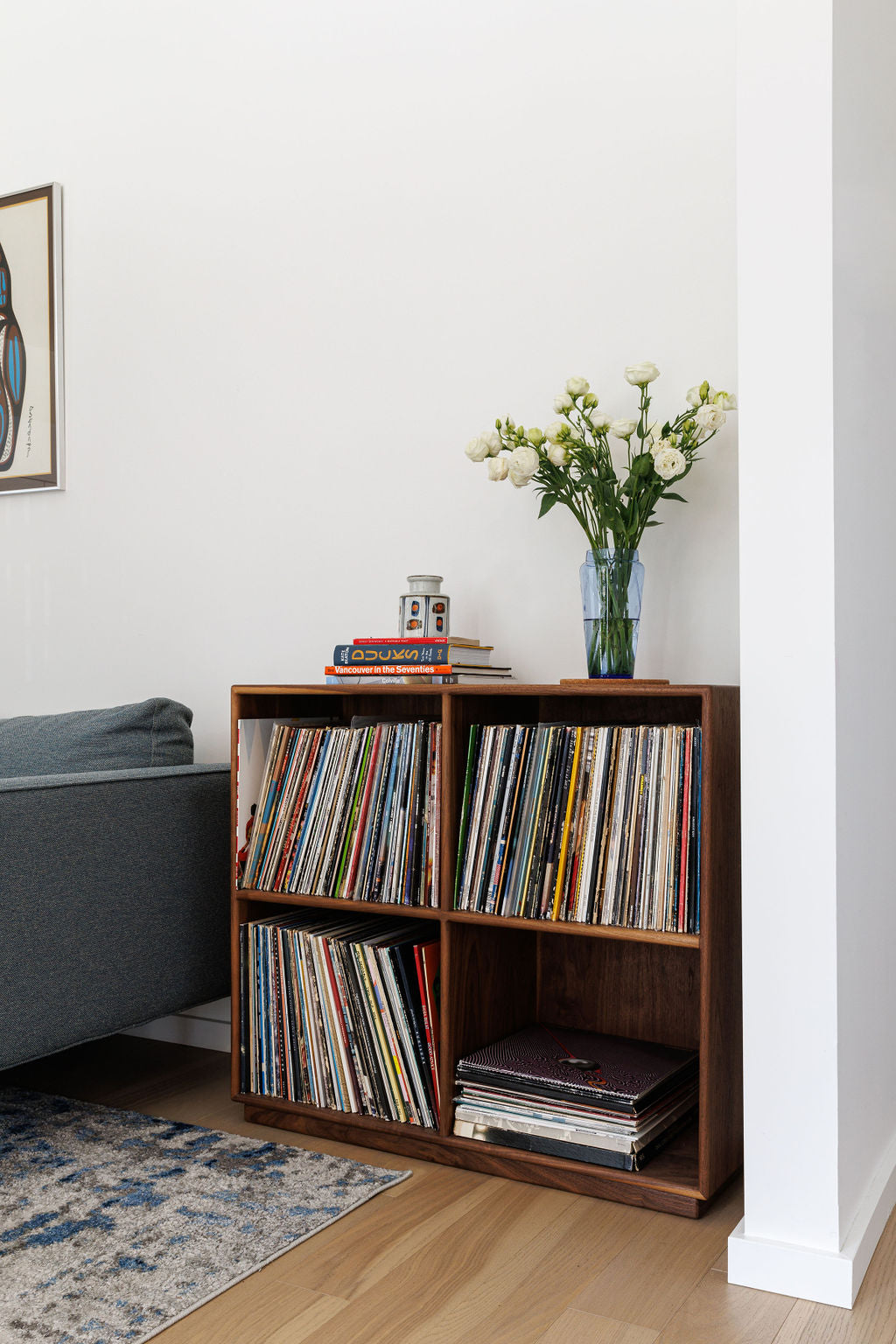 A living room scene showcasing a custom made dark wood open shelf with 4 square grid compartments. Each shelf is filled with vinyl records and a vase of flowers is placed on top.