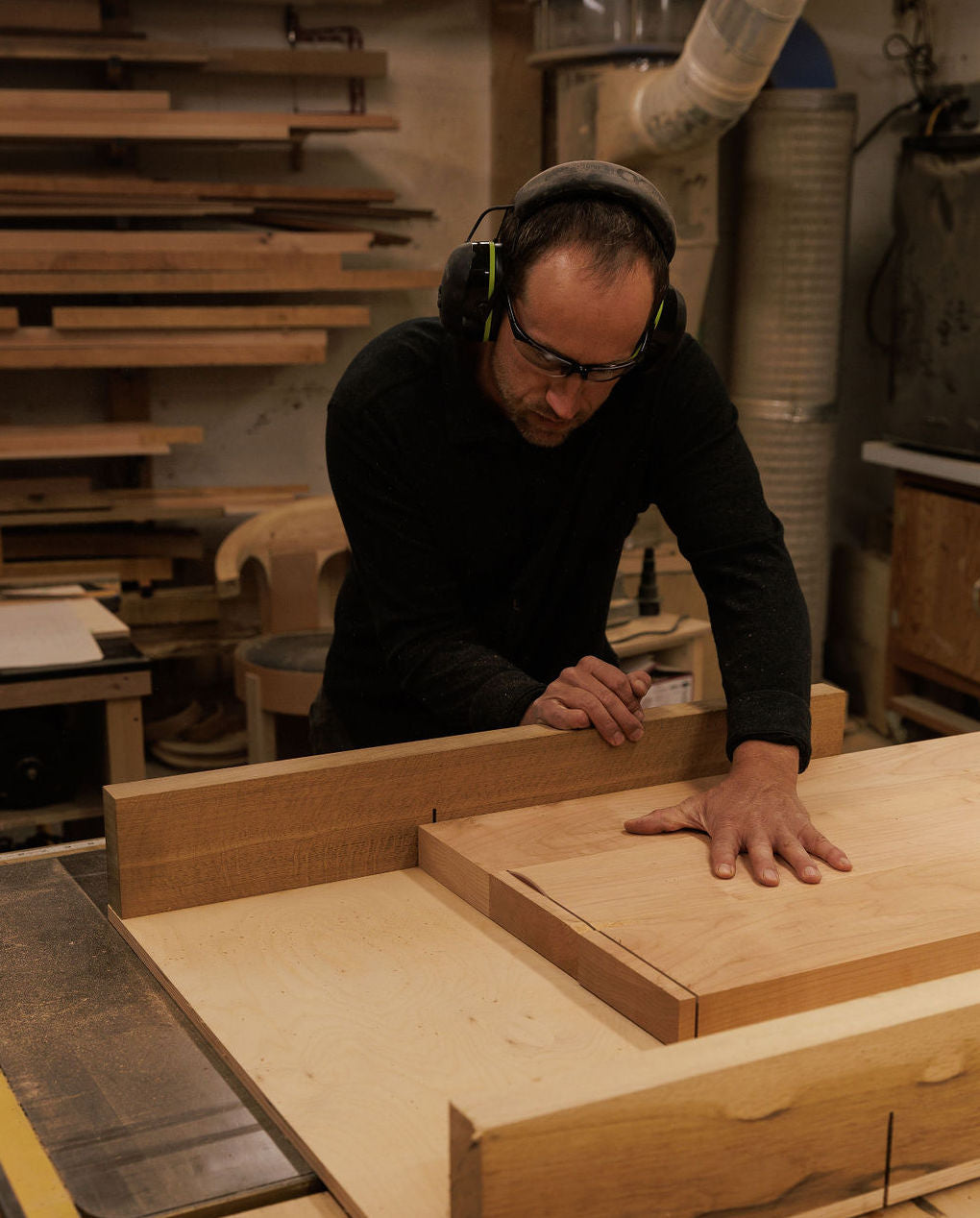 Eric is shown in his woodshop. He is wearing protective glasses and safety ear muffs while pushing a large piece of light wood through a table saw.