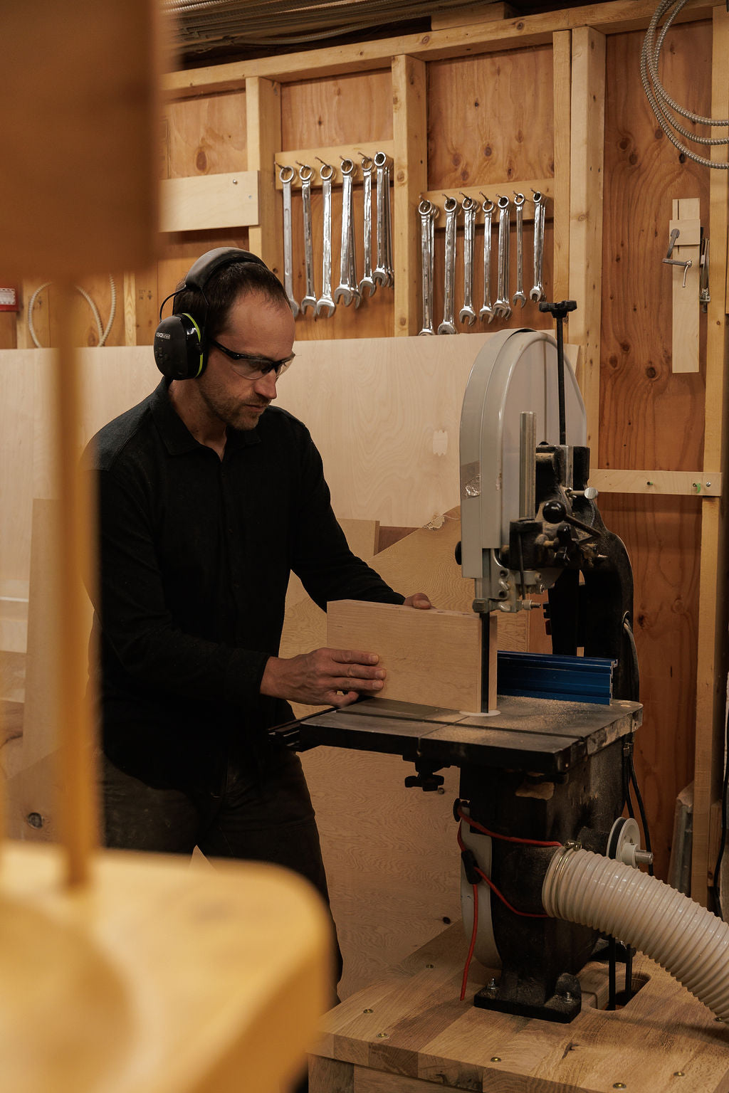 Eric stands in his wood shop at a band saw wearing protective glasses and ear muffs and guides a piece of wood through the saw.