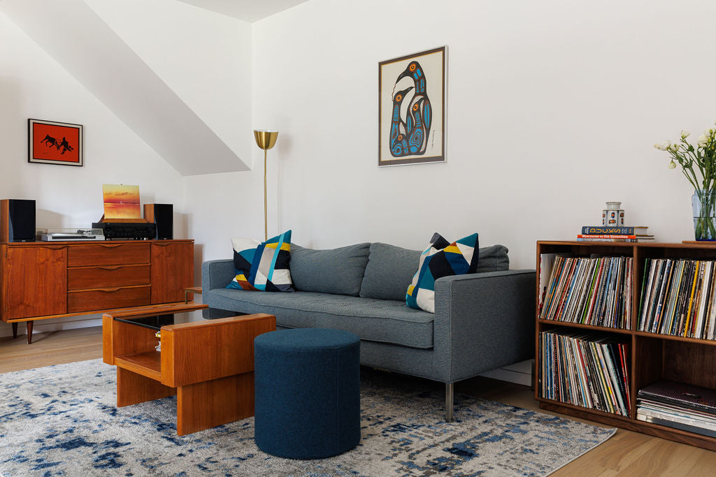 A living room scene is shown with a blue sofa. Around it are three different wooden pieces of all the same wood tone and style including a credenza, a coffee table, and a custom made free-standing cube shelf that is filled with vinyl records.
