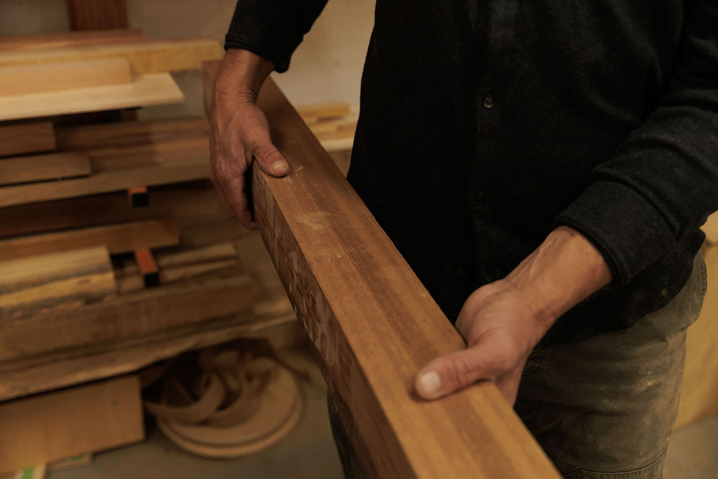 A man is carrying a piece of medium toned wood. Focused on his hands and the detail of the wood grain.