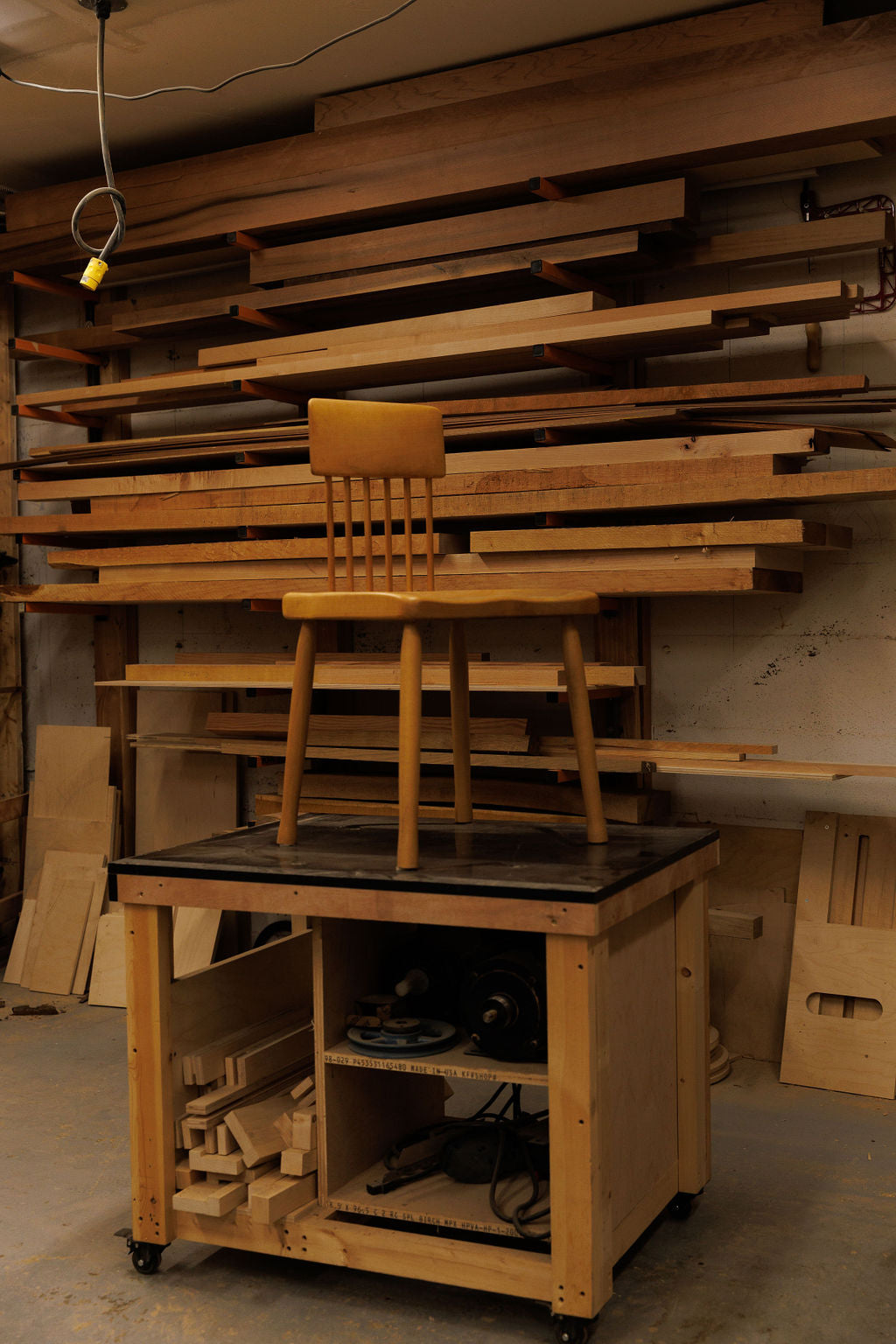 A wooden dining chair is placed on a counter in a wood shop surrounded by wood of the same warm tone.