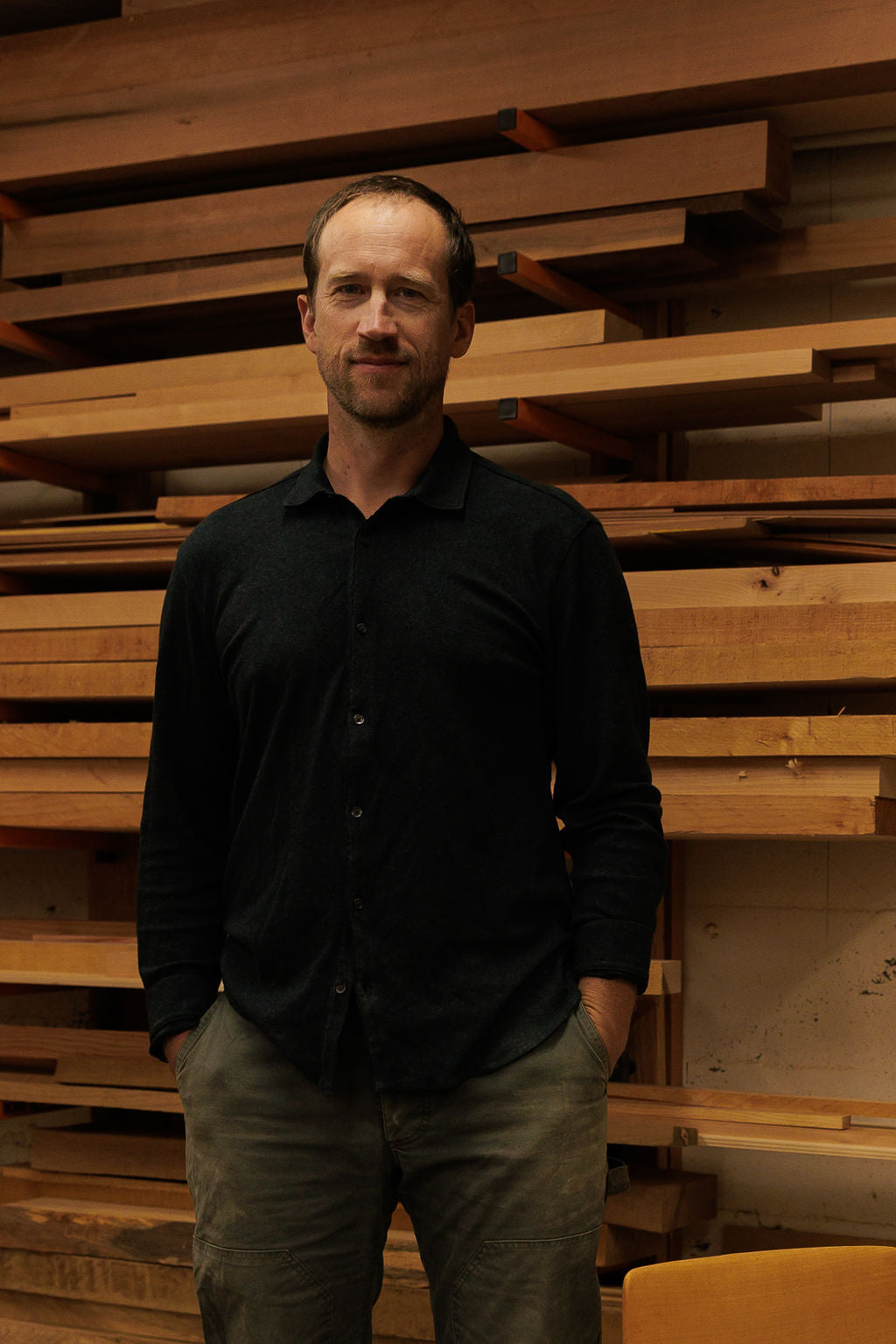 Eric stands in his wood shop in front of a shelf filled with planks of wood. He stands casually with his hands in his pockets wearing a black button up shirt. He is smiling and facing the camera.