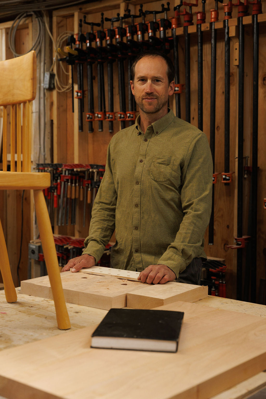 Eric is standing in his wood shop at a table where a dining room chair is placed that he is working on. He stands is front of a wall of his tools. He is smiling looking at the camera wearing a green button up shirt.